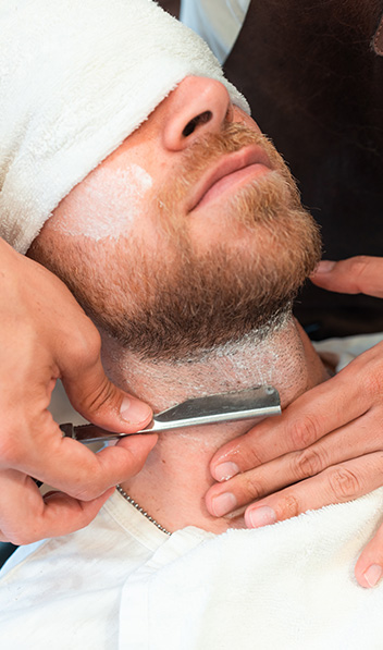 vertical-shot-hairdresser-shaving-beard-young-customer-towel-with-blade